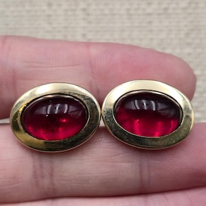 May include: A pair of oval-shaped cufflinks featuring a polished gold-tone metal frame surrounding a smooth, deep red cabochon stone. The design is symmetrical, with each cufflink displaying a rounded, translucent red center set in gold.