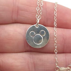 May include: A silver necklace with a round pendant featuring a Mickey Mouse silhouette. The chain is a delicate, linked style.