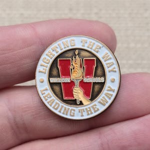 May include: A circular enamel pin featuring a white border with the text LIGHTING THE WAY LEADING THE WAY. The center displays a red letter W behind a hand holding a lit torch, with the words WILLCOX SCHOOLS printed in small black letters.