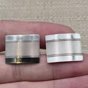 May include: A pair of rectangular silver-toned cufflinks featuring a central textured band with fine horizontal lines, bordered by smooth, polished silver-toned edges on the top and bottom, creating a clean and structured metallic appearance.
