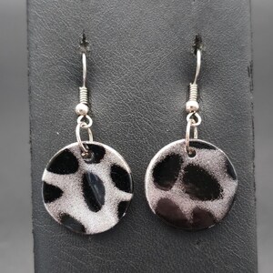 May include: A pair of dangle earrings featuring circular charms with a black and silver leopard print pattern. The earrings are attached to silver-toned metal fishhook wires, creating a simple and distinct look with a high-contrast animal motif.
