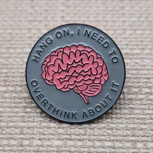 May include: A round enamel pin with a dark gray background featuring a pink anatomical brain illustration in the center. The text HANG ON, I NEED TO THINK ABOUT IT is printed in dark gray capital letters around the circular border.