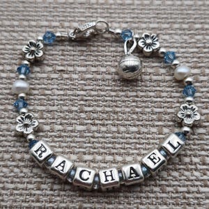 May include: A silver bracelet with blue and white beads, a silver charm, and the name "RACHEL" spelled out in silver letter beads.