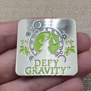 May include: A square silver-toned metal pin featuring a lime green circular center with a silhouette of a figure holding a trumpet. The design includes clock numerals, small gears, and winged figures. The text DEFY GRAVITY is printed in lime green.