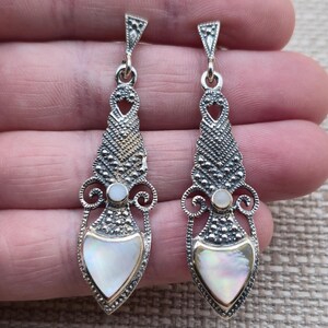 May include: A pair of silver-toned drop earrings. Each earring features a pearlescent, teardrop-shaped inlay, accented with a gold-toned border. The design includes intricate detailing and a small, round pearl-like element.