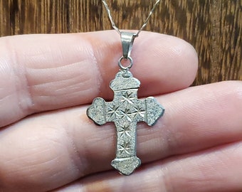 May include: Silver-colored cross pendant necklace. The cross has a textured surface with star-like patterns. The necklace chain is thin and silver, and the background is a wooden surface.