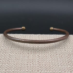 May include: A copper-colored cuff bracelet with small, gold-colored ball accents at each open end. The bracelet is displayed on a textured, light beige surface, with a dark background.