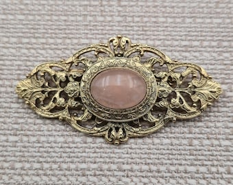 May include: An oval-shaped brooch featuring an ornate, antique gold-toned metal frame with intricate filigree scrollwork. A smooth, polished, pale pink cabochon stone is set securely in the center of the decorative metalwork design.