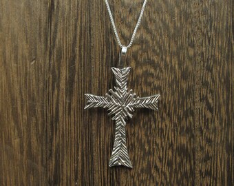 May include: A silver cross pendant with a textured, geometric design. The pendant is hanging from a silver chain.