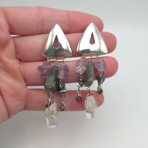 May include: Silver triangle earrings with dangling gemstones. Each earring features a silver triangle top with a cutout, from which hang various gemstones in shades of purple, green, and clear. The gemstones are wire-wrapped and suspended from delicate chains.