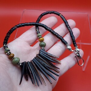 May include: A handcrafted necklace featuring a dark brown, rectangular bead chain with a pendant of dark brown, pointed, fan-shaped elements. The necklace includes green and tan spherical beads, and silver-toned accents.