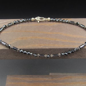 May include: Ankle bracelet featuring faceted black beads and cylindrical silver-tone metal beads. The bracelet is secured with a silver-tone clasp. The jewelry is displayed on a wooden surface.