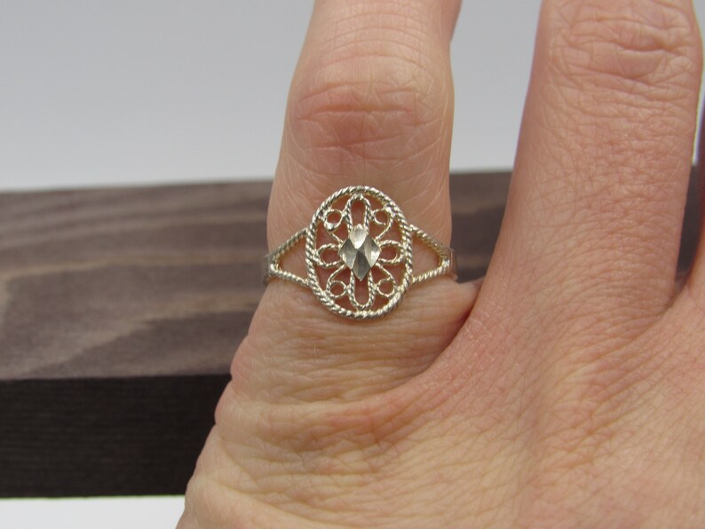 May include: A silver filigree ring with a diamond-shaped center stone. The ring is on a finger.