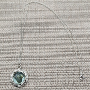 May include: A silver-toned necklace with a round pendant. The pendant features a light blue and green stone set within a textured, circular frame. The chain is delicate and has a lobster clasp closure.