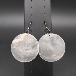 May include: A pair of circular drop earrings crafted from thin, translucent, off-white shell material. Each earring features a smooth, flat, round disc with natural swirling patterns and subtle iridescent tones, suspended from simple silver-toned metal hooks.