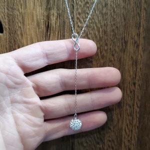 May include: A silver-colored necklace featuring an infinity symbol and a dangling chain. The chain ends with a round, sparkling, crystal-covered ball. The necklace is displayed against a wooden background.