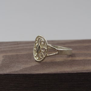 May include: A silver ring with an oval-shaped filigree design. The ring is on a brown wooden surface.