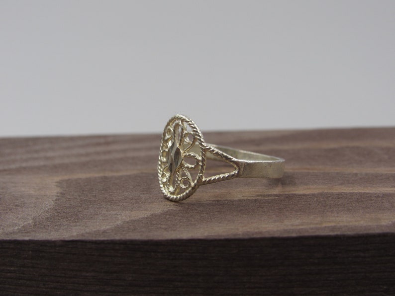 May include: A silver ring with an oval-shaped filigree design. The ring is on a brown wooden surface.