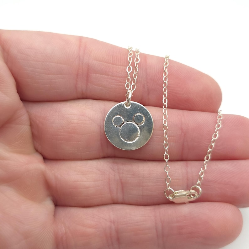 May include: A silver necklace with a round pendant featuring a Mickey Mouse silhouette. The chain is a delicate, linked style.
