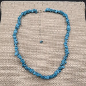 May include: A turquoise chip necklace with a silver-colored chain and clasp. The necklace is composed of small, irregularly shaped turquoise stones, strung together to form a choker-style necklace. The background is a neutral tan fabric.