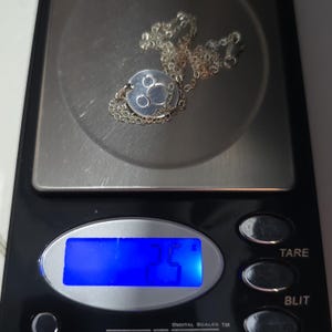 May include: A silver necklace with a round pendant featuring three circles. The necklace is on a digital scale that reads 2.5.