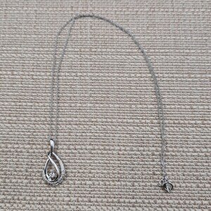 May include: A silver-colored necklace with a teardrop pendant. The pendant features a central clear stone surrounded by smaller stones and a twisted metal design. The chain is delicate and has a lobster clasp.
