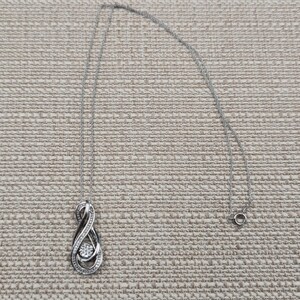 May include: A silver-toned pendant necklace with an infinity symbol design. The pendant features a central, round, clear gemstone surrounded by smaller stones. The chain is delicate and the clasp is visible.