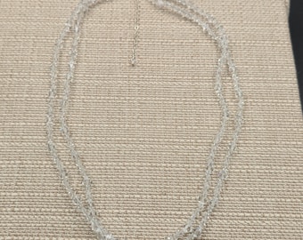 May include: A clear, double-strand necklace with small, faceted beads. The necklace has a delicate, shimmering appearance and a silver-toned clasp. The beads are evenly spaced, creating a symmetrical design. The necklace is displayed on a neutral-toned fabric.