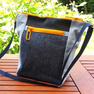 Op de afbeelding: Een blauwe denim crossbody tas met een gele rits en een blauwe band. De tas ligt op een houten tafel.