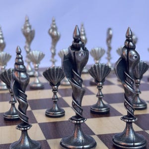 May include: A chess set featuring silver-coloured pieces with detailed designs. The pieces include spiral and fan shapes, arranged on a checkered wooden board. The chess pieces have an antique finish and intricate detailing.