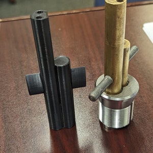 May include: Two metal parts, one black and one silver, with a brass tube attached to the silver part. The black part has a cross-shaped base and two holes at the top. The silver part has a threaded base and a cross-shaped handle.