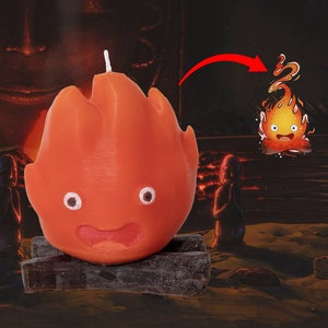 Puede incluir: Vela naranja con forma de espíritu de fuego de la película "Mi vecino Totoro". La vela tiene una cara sonriente y dos ojos grandes. La vela está sentada sobre una base negra que parece troncos.