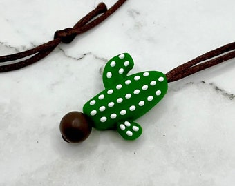 May include: A green cactus charm with white dots hangs from a brown cord necklace. The charm is made of polymer clay and has a brown bead at the top.