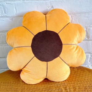 Puede incluir: Una almohada de felpa con forma de girasol con un centro marrón y pétalos amarillos. La almohada está sentada sobre una superficie marrón texturizada.