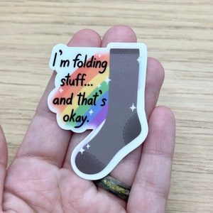 以下が含まれることがあります： グレーの靴下と虹を背景にしたステッカー。ステッカーのテキストは「私は物を畳んでいます…そしてそれは大丈夫なのです。」