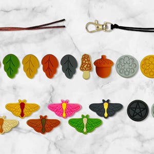 Puede incluir: Una colección de dijes decorativos y llaveros en varias formas y colores. Incluye diseños de hojas, hongos, bellotas y mariposas, junto con dijes circulares y ovalados con patrones de estrellas y geométricos. También hay dos llaveros con cierres dorados.