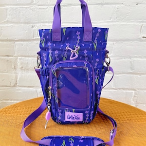 May include: A purple tote bag with a floral pattern of lavender sprigs. The bag has a clear pocket, a key charm, and a detachable shoulder strap. The bag has a top handle and a logo that says "Artie Xan".