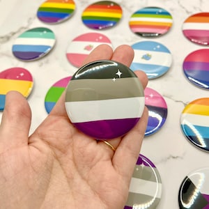 May include: A collection of colorful pride flag pins, including a gray, white, and purple asexual pride flag pin.