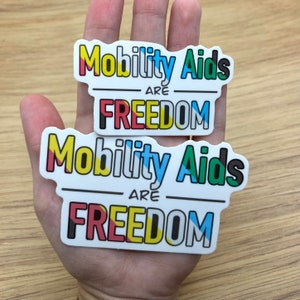 May include: Two white stickers with the text "Mobility Aids Are Freedom" in colourful letters. The stickers are layered on top of each other.