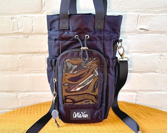 May include: A navy blue nylon crossbody bag featuring a drawstring top closure, two top handles, and an adjustable shoulder strap. The front includes a rectangular clear plastic pocket and a black patch with the white text ArtisXan.