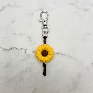 May include: A yellow sunflower keychain with a brown center and a brown cord attached to a silver key ring.