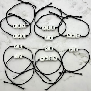 May include: Ten black string bracelets with white letter beads spelling out the words "FEY", "FAE", "ZIE", "ZE", "SHE", "VE", "EY", "XE", "THEY", and "HE".
