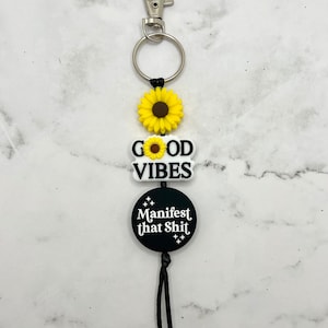 May include: A black and white keychain with a sunflower charm, a charm that says "GOOD VIBES" with a sunflower in the middle, and a round charm that says "Manifest that Shit" with stars around the text. The keychain has a black cord and a silver key ring.
