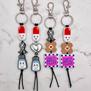 May include: Four colourful keychains with different charms. The charms include a pill, a stethoscope, a bandage, and a salt shaker. The keychains also have black tassels and the words "Back & Body Hurts" on the bottom.