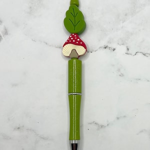 May include: A green pen with a mushroom and leaf design. The mushroom is red with white dots and the leaf is green.