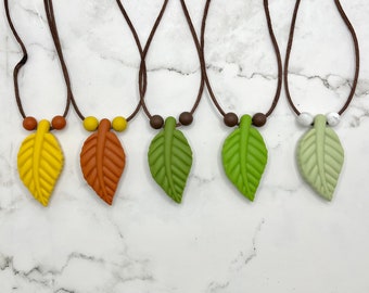 May include: Five necklaces with leaf pendants in different colors. The necklaces are brown cords with brown beads and leaf pendants in yellow, orange, green, green, and light green.