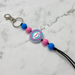 Peut inclure: Un porte-clés blanc avec un cercle bleu et rose qui dit "Protéger les droits des trans". Le porte-clés a un anneau de clé argenté et un cordon noir avec des perles bleues et roses.