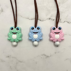 May include: Three silicone frog necklaces with brown cords. The frogs are green, blue, and pink with white eyes and white bellies. Each frog has a white bead at the bottom of the cord.