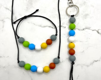 May include: A set of three necklaces with colorful silicone beads. The necklaces are made of black cord and have a variety of colors, including gray, green, blue, white, and yellow. The longest necklace has a silver key ring attached to it.