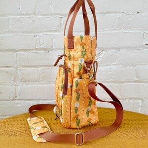 Op de afbeelding: Een kleine, gele en bruine tote bag met een bloemenprint. De tas heeft een bovenste handvat en een lange verstelbare schouderband. De tas heeft een ritsvak aan de voorkant en een klein vak aan de zijkant.